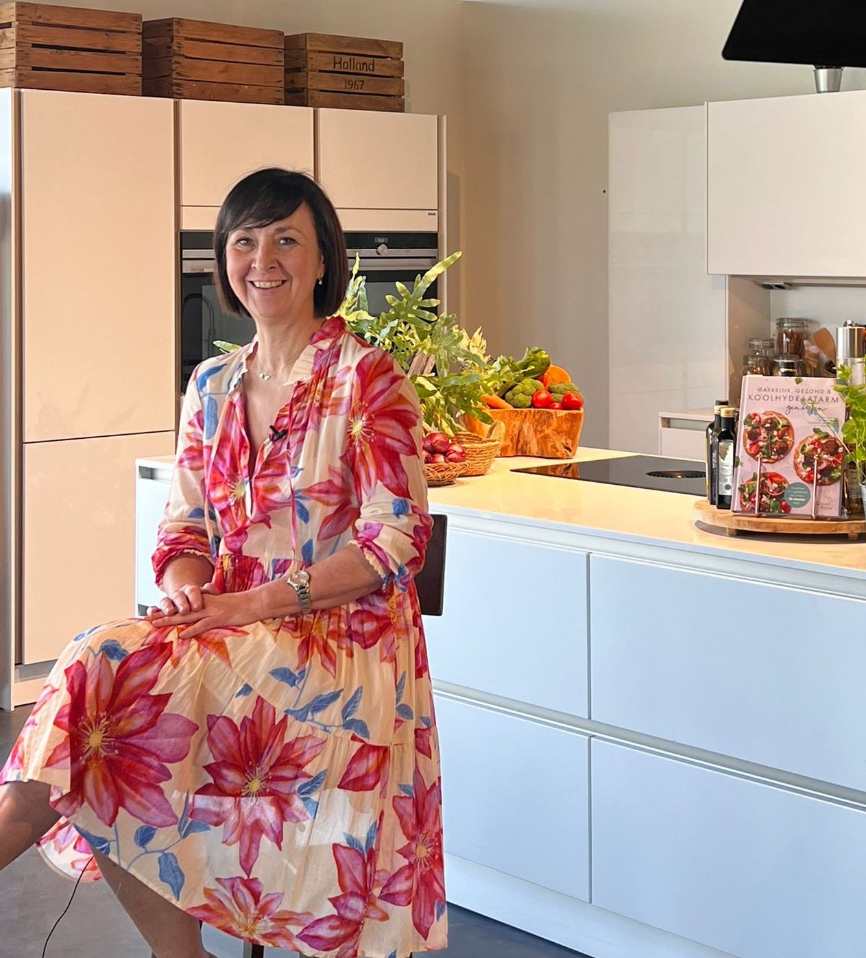 Eigenaresse van salon Isabelle in moderne keuken, zittend in kleurrijke bloemenjurk met verse groenten op het aanrecht.